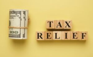 A roll of hundred-dollar bills next to wooden blocks spelling "TAX RELIEF," representing New Jersey property tax savings for retirees through the ANCHOR, Senior Freeze, and Stay NJ programs.