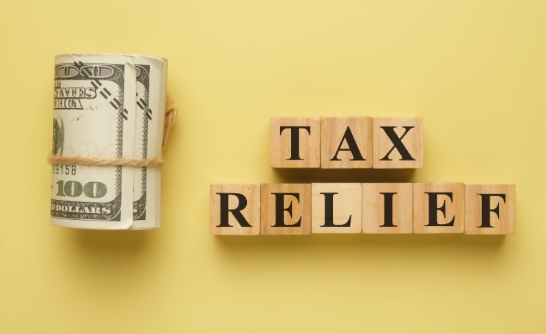 A roll of hundred-dollar bills next to wooden blocks spelling "TAX RELIEF," representing New Jersey property tax savings for retirees through the ANCHOR, Senior Freeze, and Stay NJ programs.