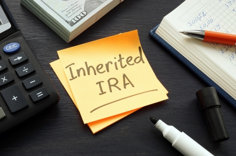 An orange sticky note reading "Inherited IRA" surrounded by a calculator, cash, and a financial notebook on a dark desk.